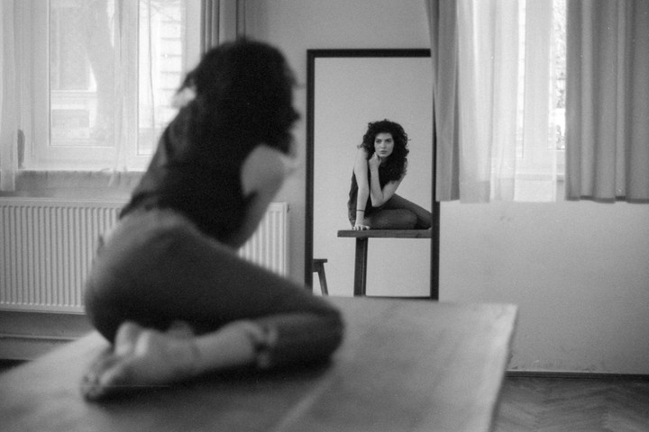 A woman kneels on a table and looks at her reflection in a mirror.