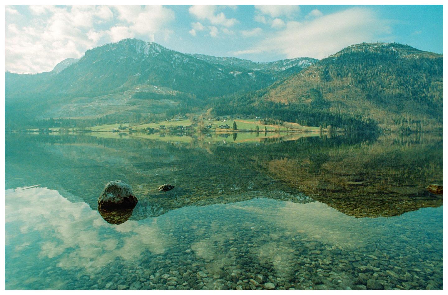 A calm lake reflects mountains and a village under a cloudy sky.