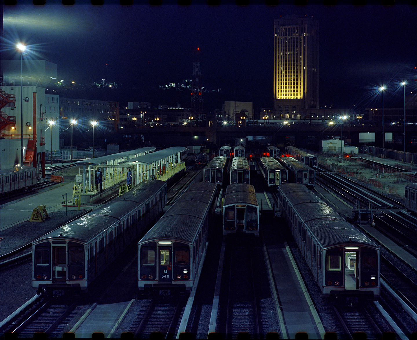 A rail yard at night is filled with trains on tracks, with a tall building illuminated in the background.