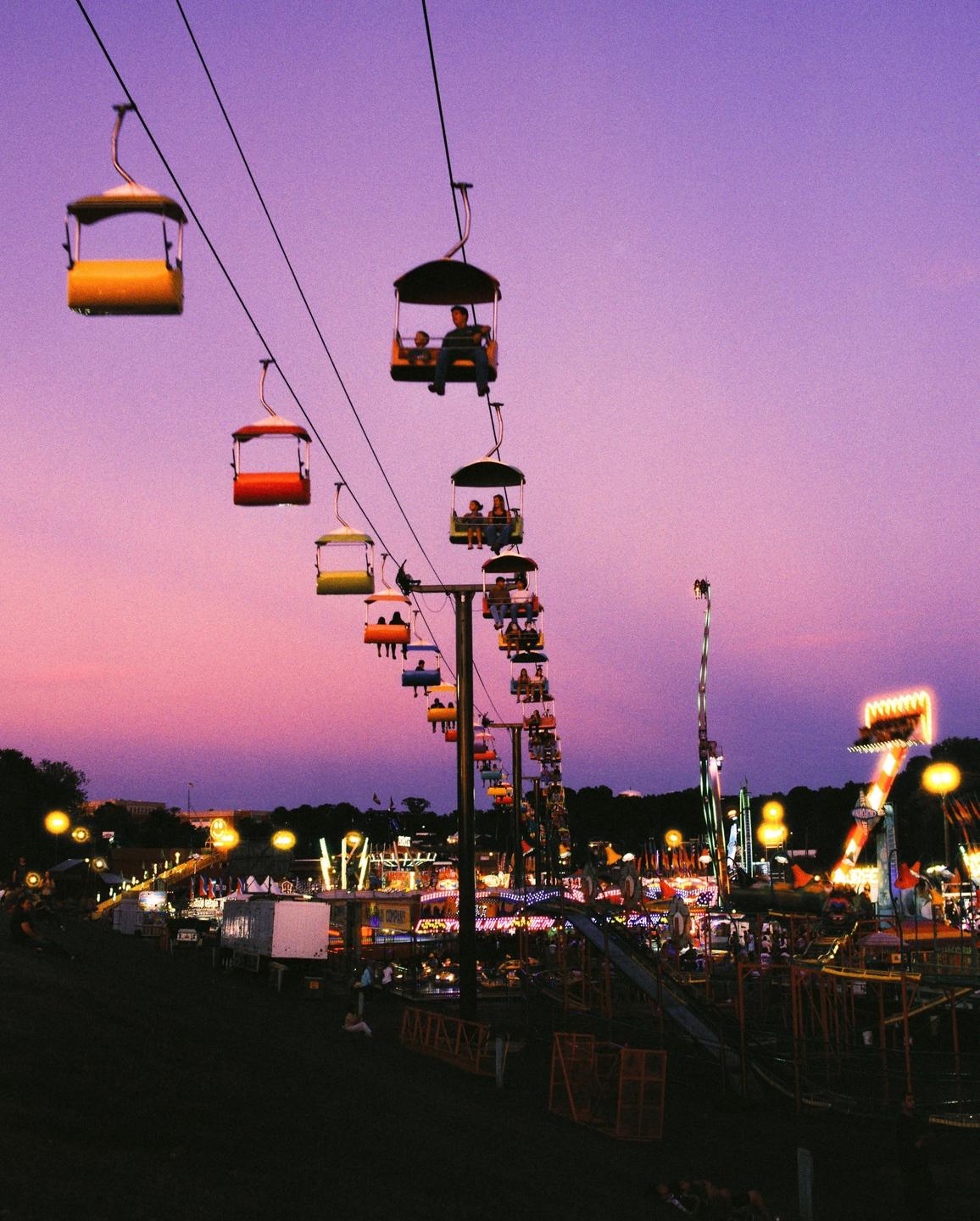 A long line of colorful gondolas with people inside travels across a fairground at dusk.