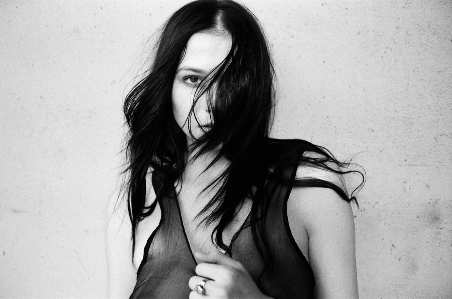 A woman with long dark hair covering part of her face wears a sheer top and a ring on her finger.