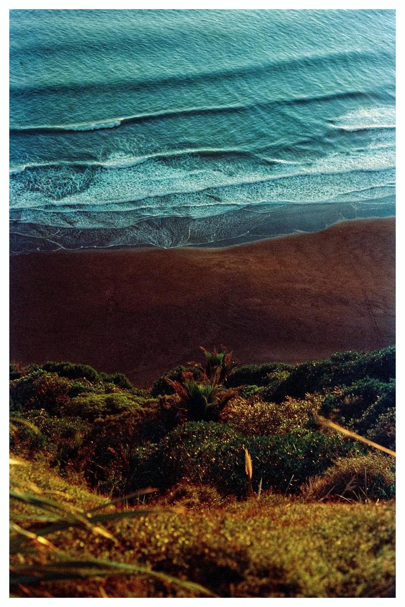 An aerial view shows waves breaking on a dark sand beach next to a lush green hillside.