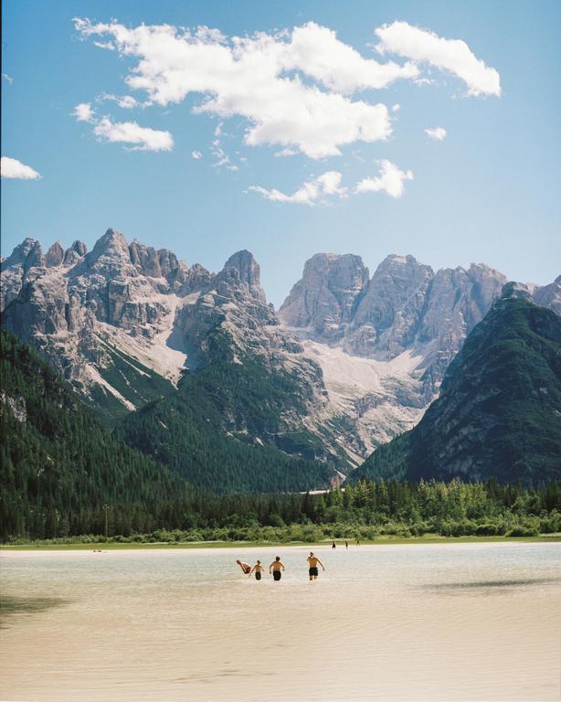 Several people wade in a shallow lake with a forest and mountains in the background under a blue sky with clouds.