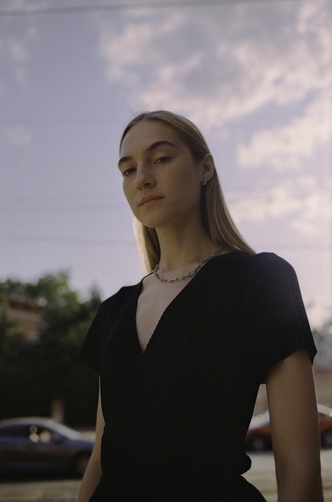 A woman with long hair and a black top looks directly at the viewer under a cloudy sky.