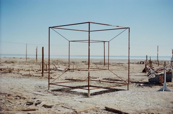 A metal cube frame stands on a dry, barren landscape with a body of water in the distance under a clear sky.