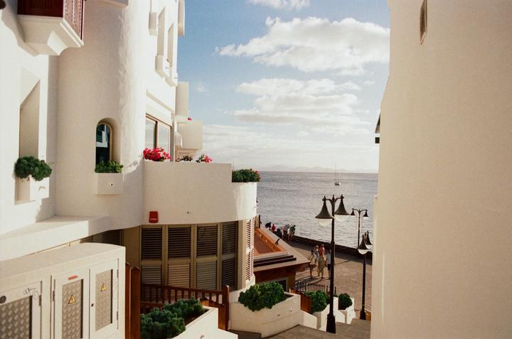 A white building with balconies and red flowers overlooks a body of water with a sailboat and distant mountains under a cloudy sky.