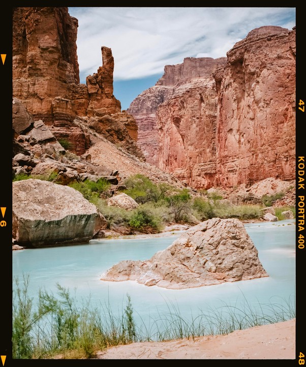 A light blue river flows through a canyon with tall red rock formations and green vegetation.