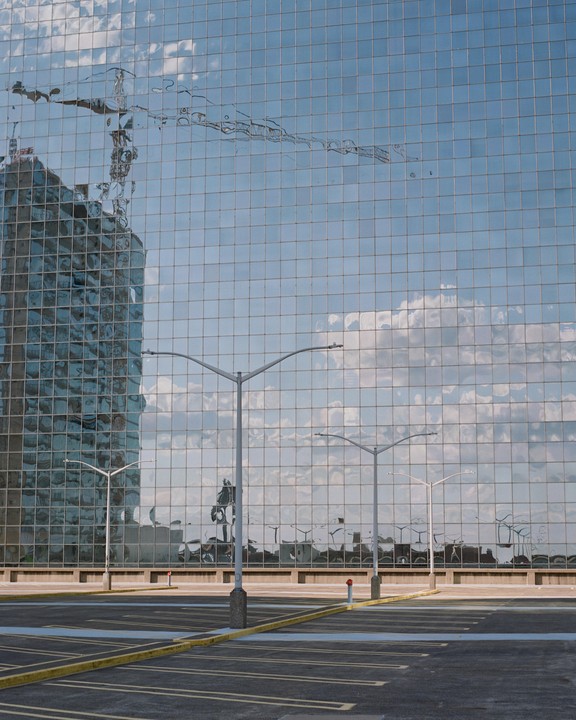 A parking lot with streetlights is in front of a glass building reflecting the sky and another building.