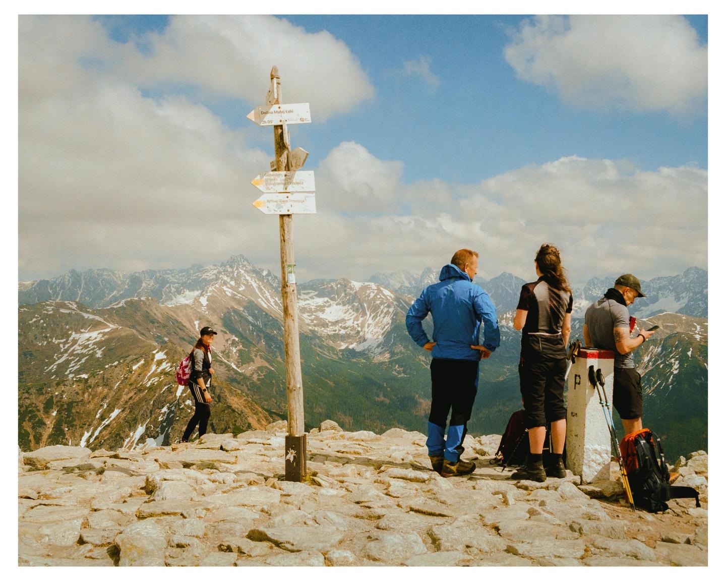 A group of hikers stands on a rocky mountain peak with snow-capped mountains in the background.