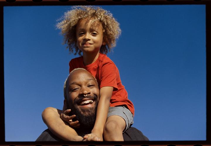 A man with a beard and a child with curly hair are smiling at the camera against a clear blue sky.
