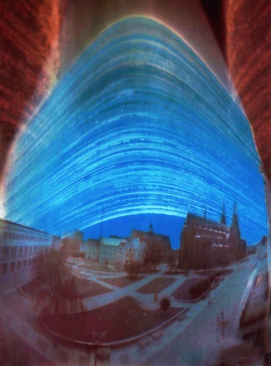 A long exposure photograph shows star trails over a town square with buildings and a church.
