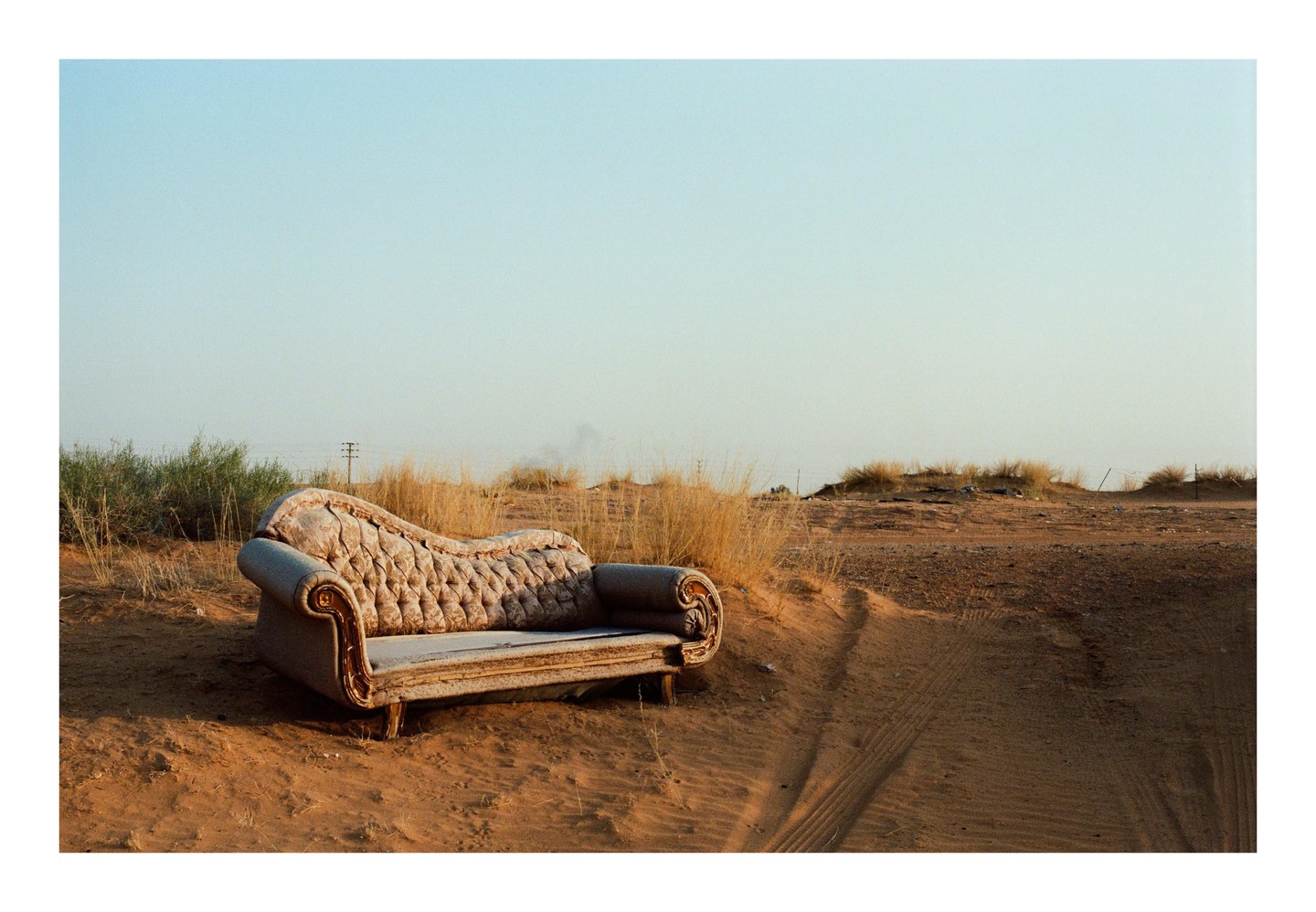 An ornate sofa sits abandoned in a sandy, arid landscape with sparse vegetation under a clear sky.