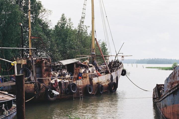 Two old, rusty boats are docked in a calm body of water next to a lush green forest under an overcast sky.