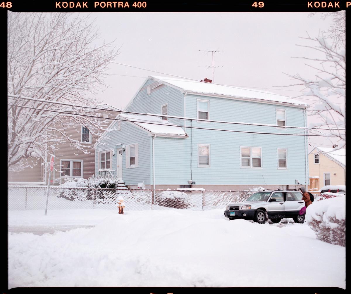 A light blue house and a silver car are covered in snow on a winter day.