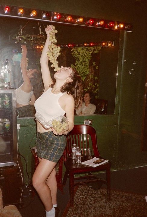 A woman in a plaid skirt eats grapes from a bunch held above her head, with another woman visible in a mirror reflection.