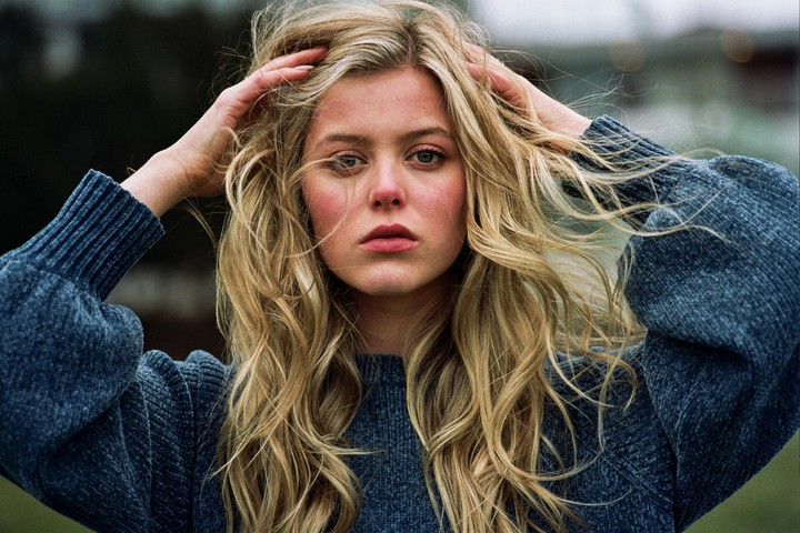 A woman with long blonde hair and a blue sweater holds her hands to her head.