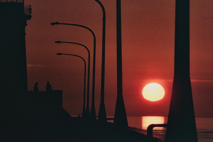 Two men stand on a pier with streetlights as the sun rises over the water.