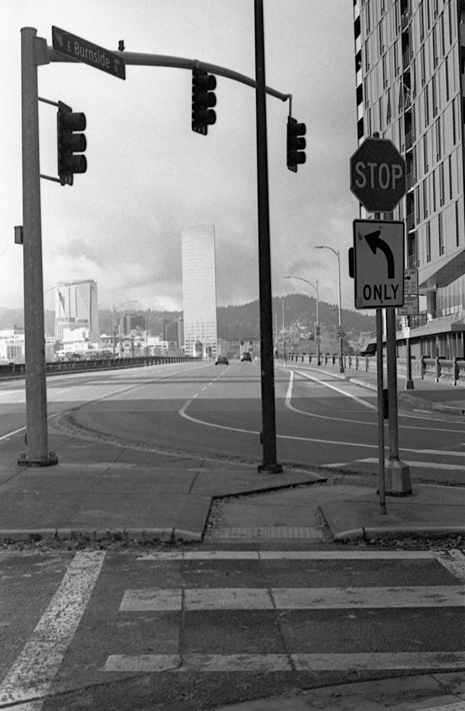 A black and white street photograph shows a city street with a stop sign and traffic lights, with buildings and hills in the background.