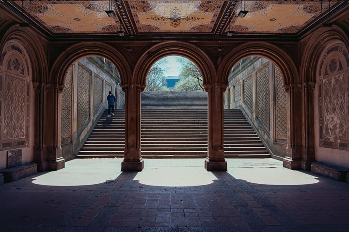 A person walks up a grand stone staircase under an ornate arched ceiling with light casting shadows on the floor.
