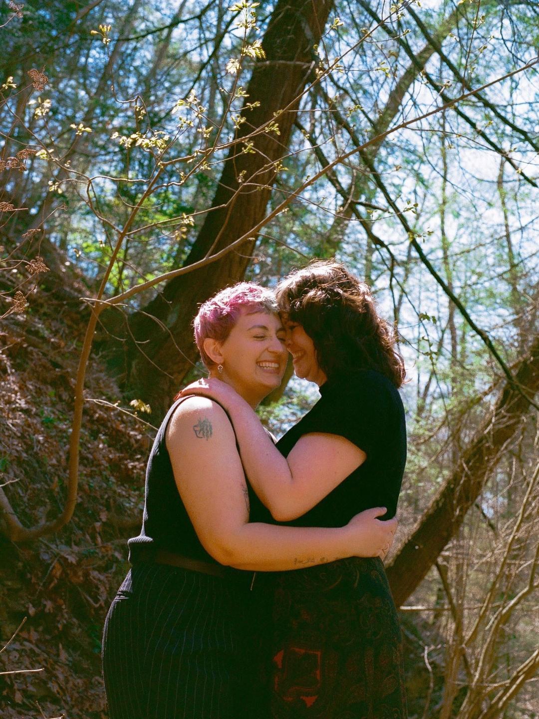 A couple embraces and smiles in a forest with dappled sunlight.