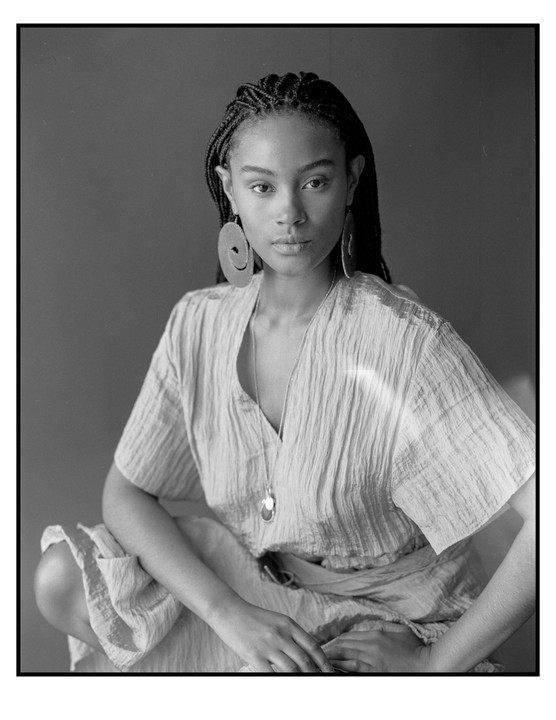 A woman with braided hair and large earrings is crouching while looking at the viewer.