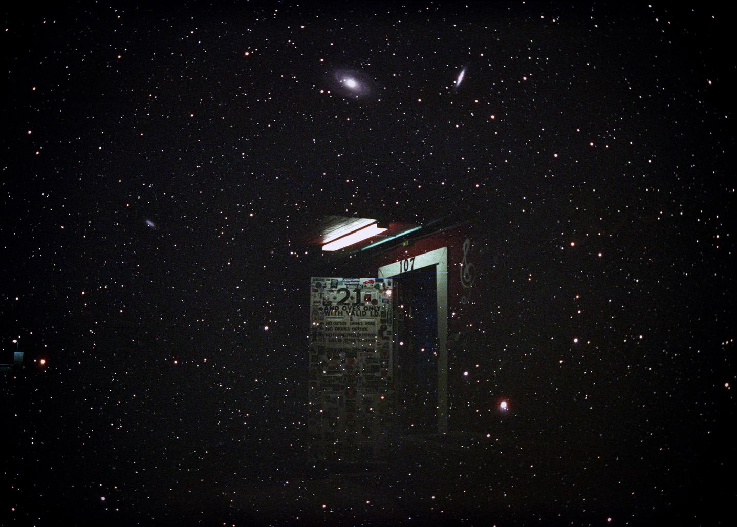 A dark night sky filled with stars and galaxies is visible above a building entrance with a sign.