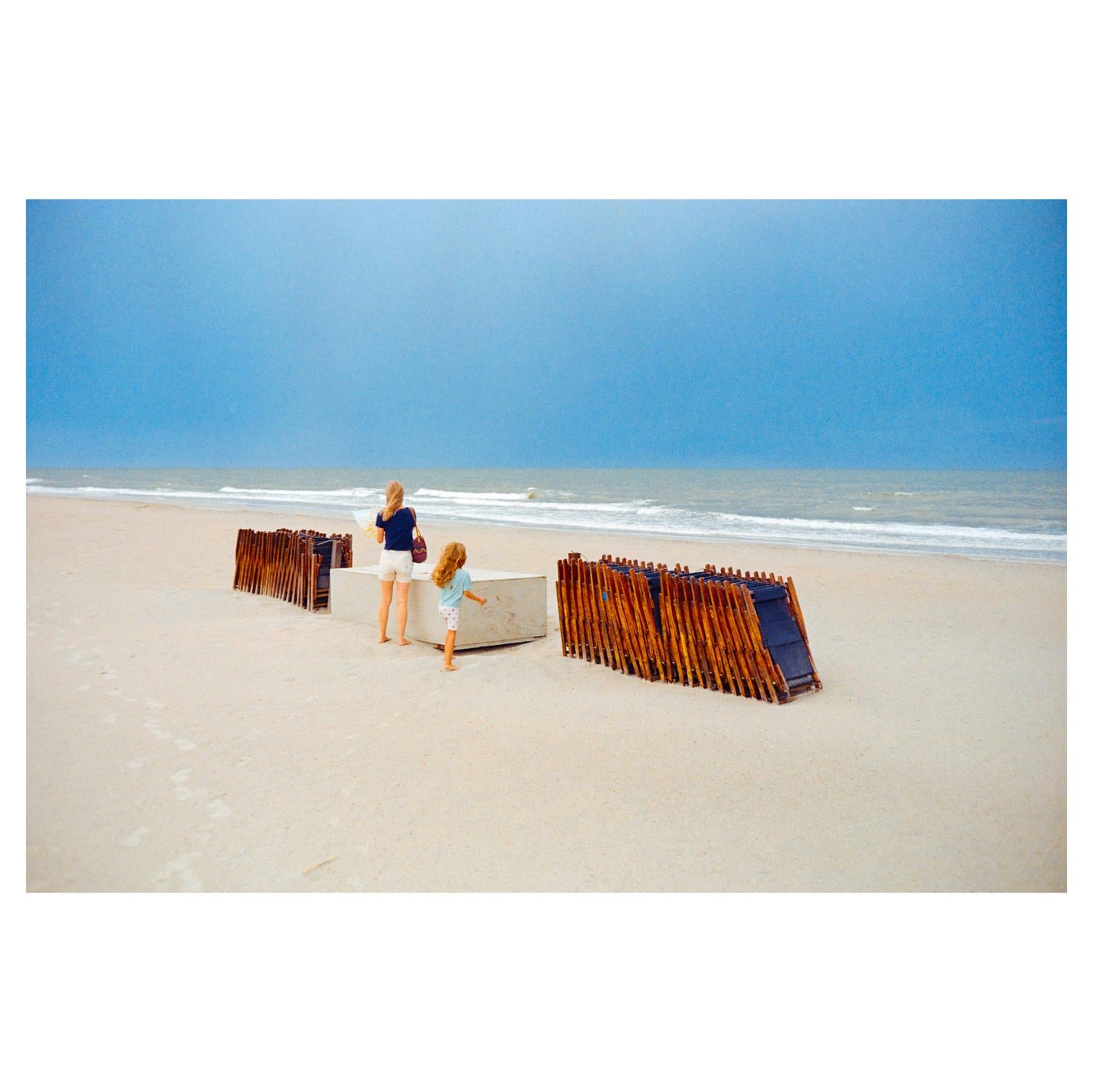 A woman and a child stand on a sandy beach next to stacked wooden beach chairs, looking out at the ocean under a blue sky.