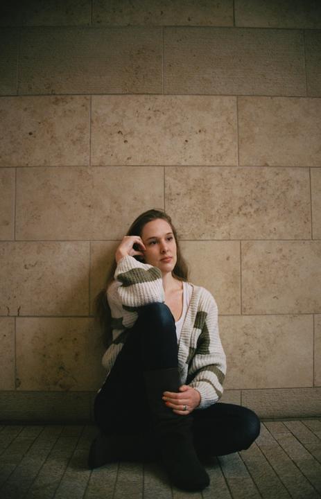 A woman sits on a tiled floor with her back against a stone wall, looking away from the camera.