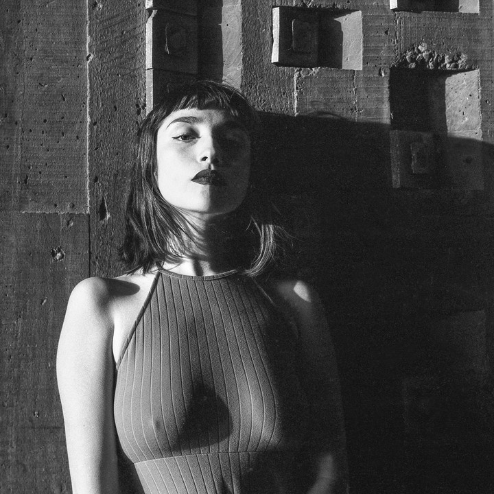A woman with dark lipstick and bangs looks directly at the viewer, leaning against a textured wall with strong shadows.