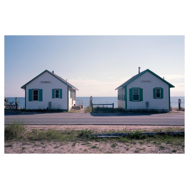 Two small white houses with green trim and shutters stand on either side of a road, with the ocean visible in the background.