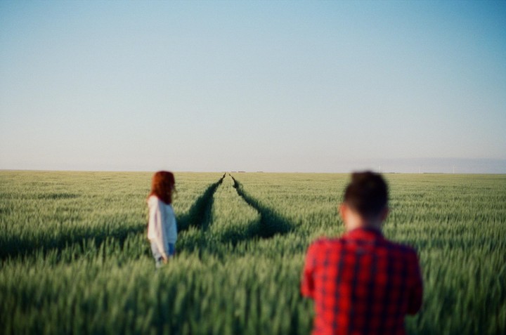 A man in a red plaid shirt stands in a field of green crops, looking towards a woman with red hair and a path leading into the distance.