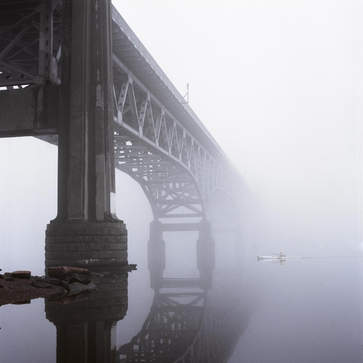 A person in a kayak paddles on a body of water next to a large bridge on a foggy day.
