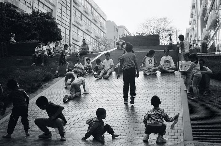 A group of children and adults are gathered on a paved incline next to a building.