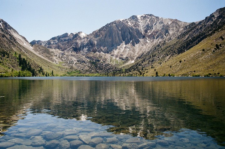 A clear lake reflects a mountain range with patches of snow and green slopes under a bright sky.