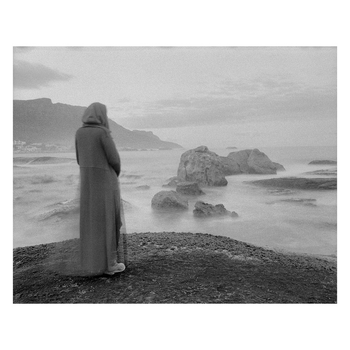 A person in a long coat stands on a rocky shore looking out at the ocean with large rocks and mountains in the distance.