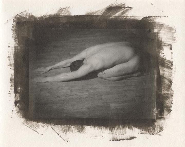 A nude man is kneeling on a wooden floor with his head down and arms stretched forward.