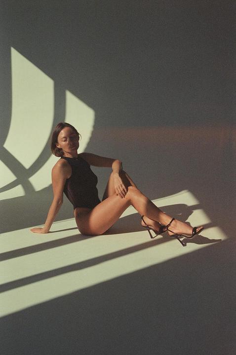 A woman in a dark top and high heels sits on the floor with strong shadows cast around her.