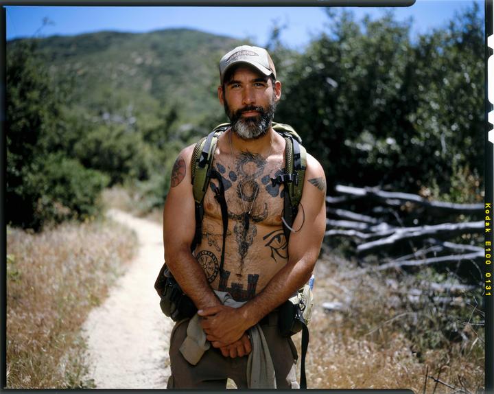 A man with a beard and tattoos stands on a dirt path with a backpack, surrounded by dry grass and green foliage.