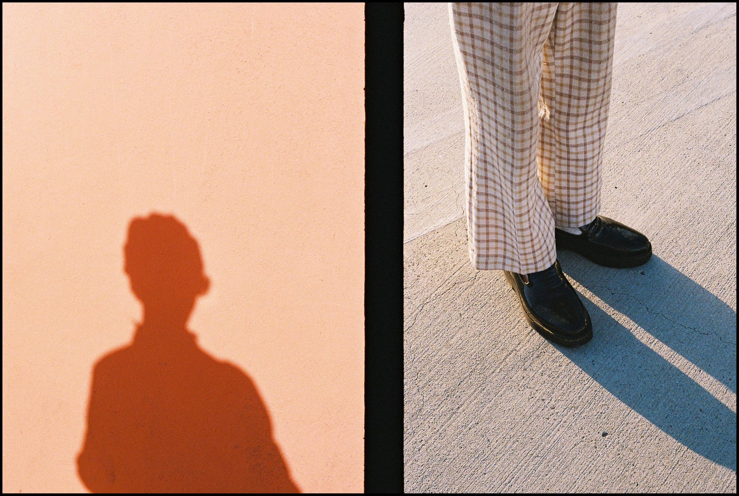 A person's shadow is cast on a peach-colored wall next to their legs and feet on a concrete surface.