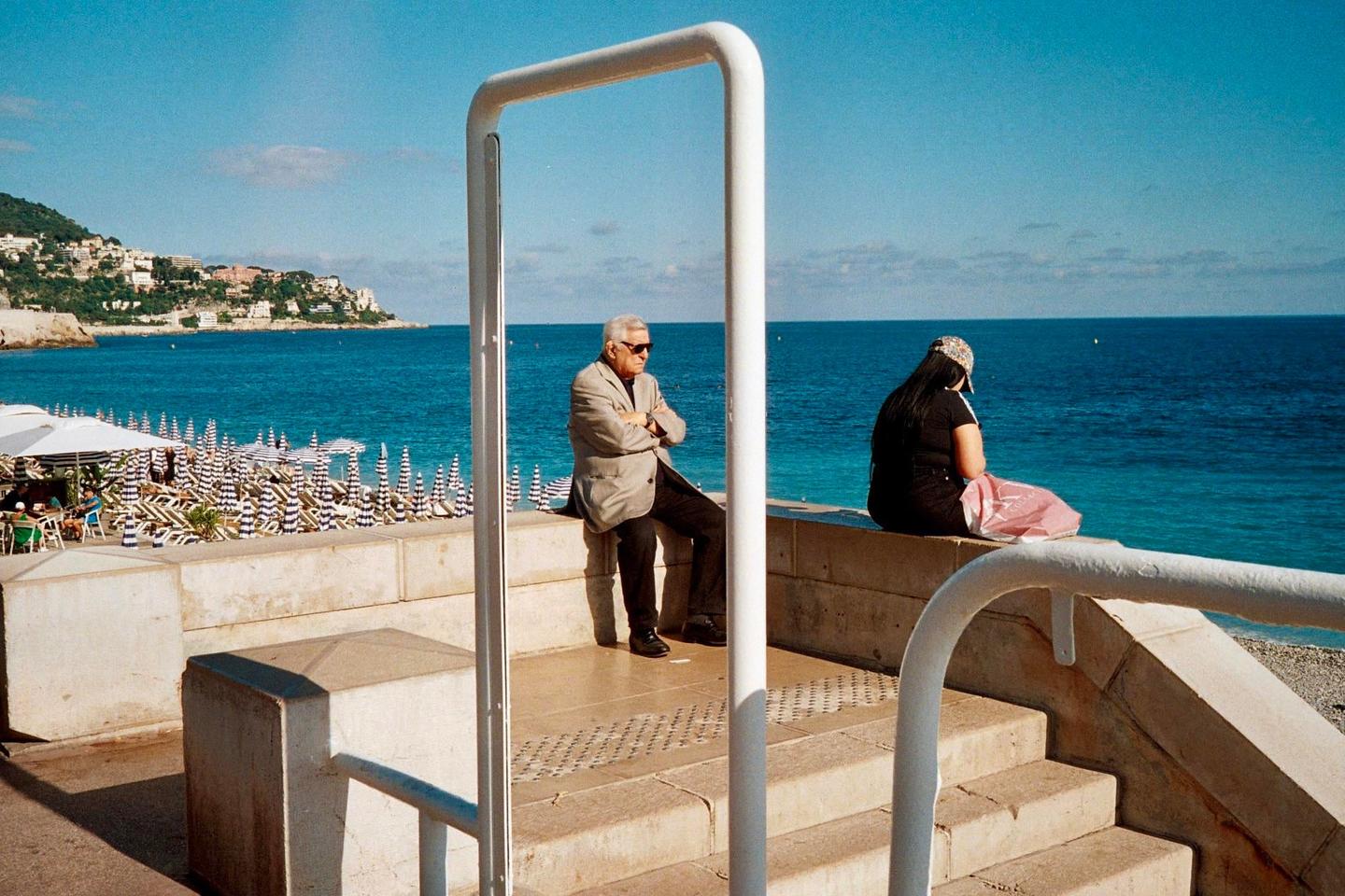 A man and a woman sit on a stone wall overlooking a beach with striped umbrellas and the ocean.