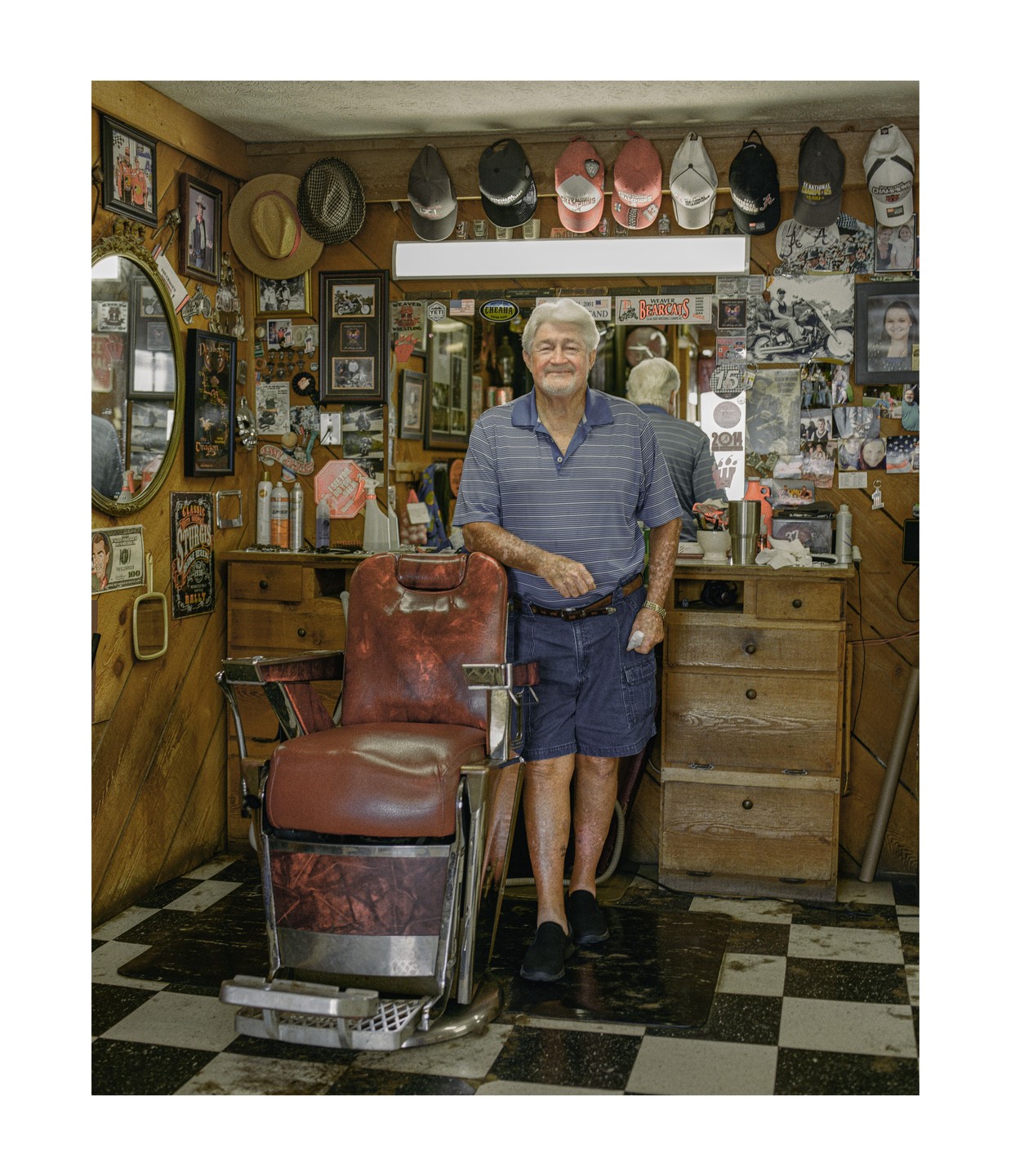An elderly man stands next to a barber chair in a cluttered barbershop.