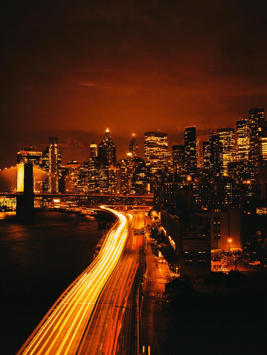 A long exposure photograph captures the light trails of vehicles on a highway next to a river, with a city skyline and bridge in the background at night.