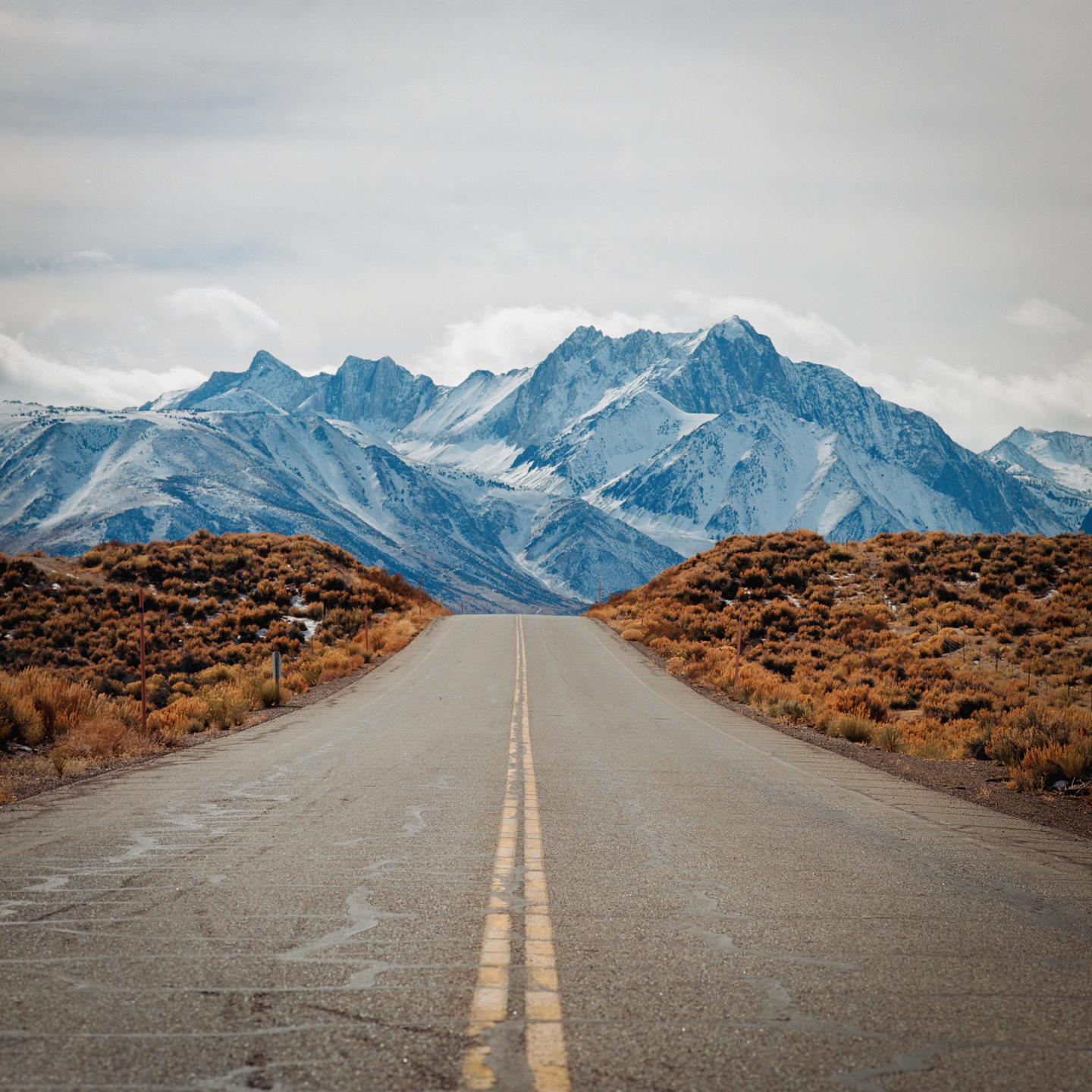A long road with double yellow lines leads towards snow-capped mountains under a cloudy sky.
