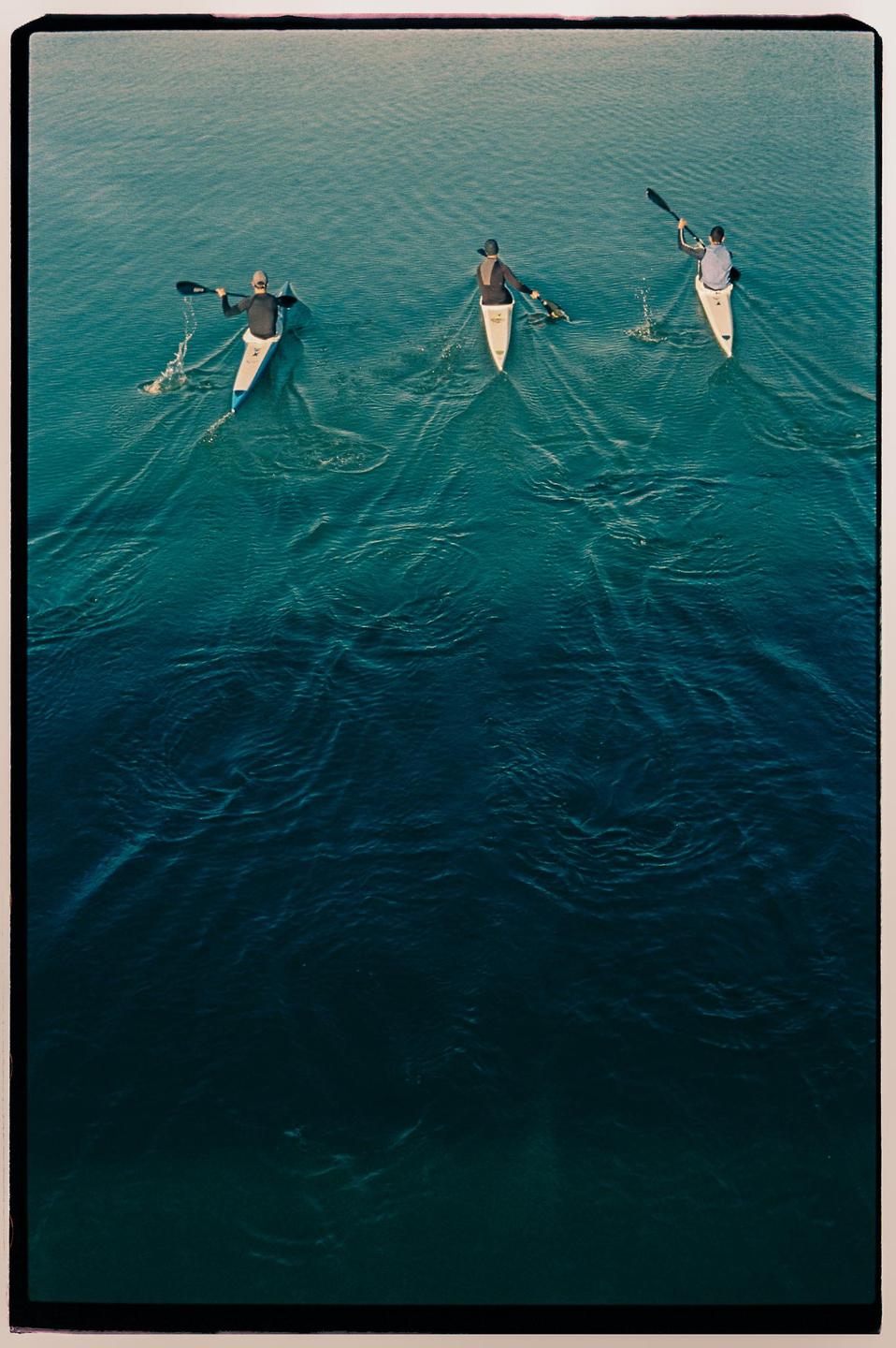 Three people in kayaks paddle across a body of water, viewed from above.