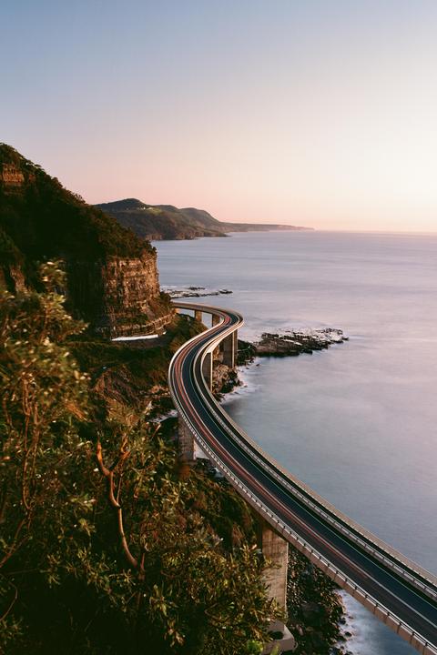 A long exposure shot captures the light trails of vehicles on the Sea Cliff Bridge as it curves along the coastline at dusk.