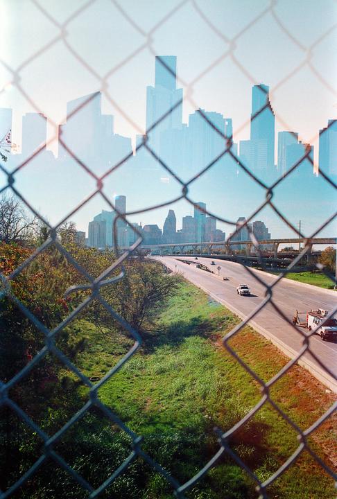 A double exposure shows a chain link fence and a highway in the foreground, with a city skyline superimposed in the background.