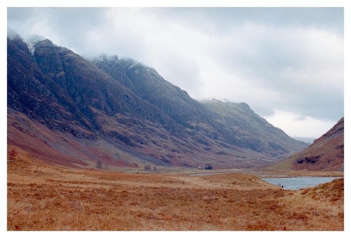 A vast valley with a lake and mountains partially covered in clouds is visible under a cloudy sky.