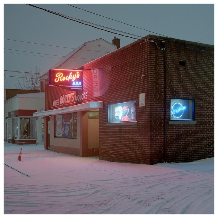 A brick building with neon signs for Rocky's Bar and Liquors stands in a snow-covered landscape at night.