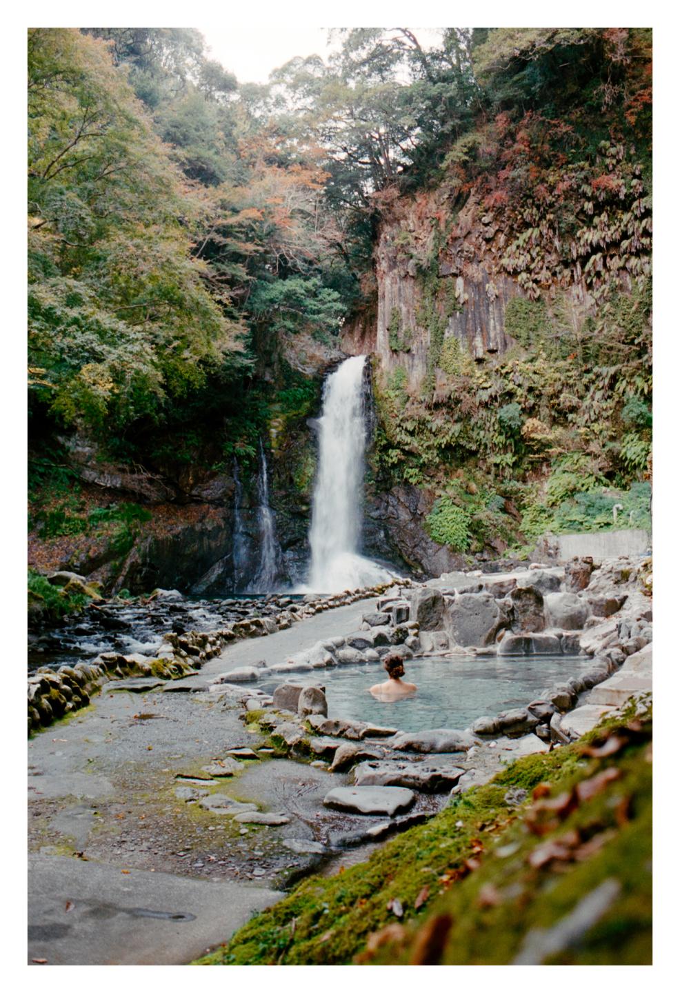 A person is bathing in an outdoor onsen with a waterfall in the background.