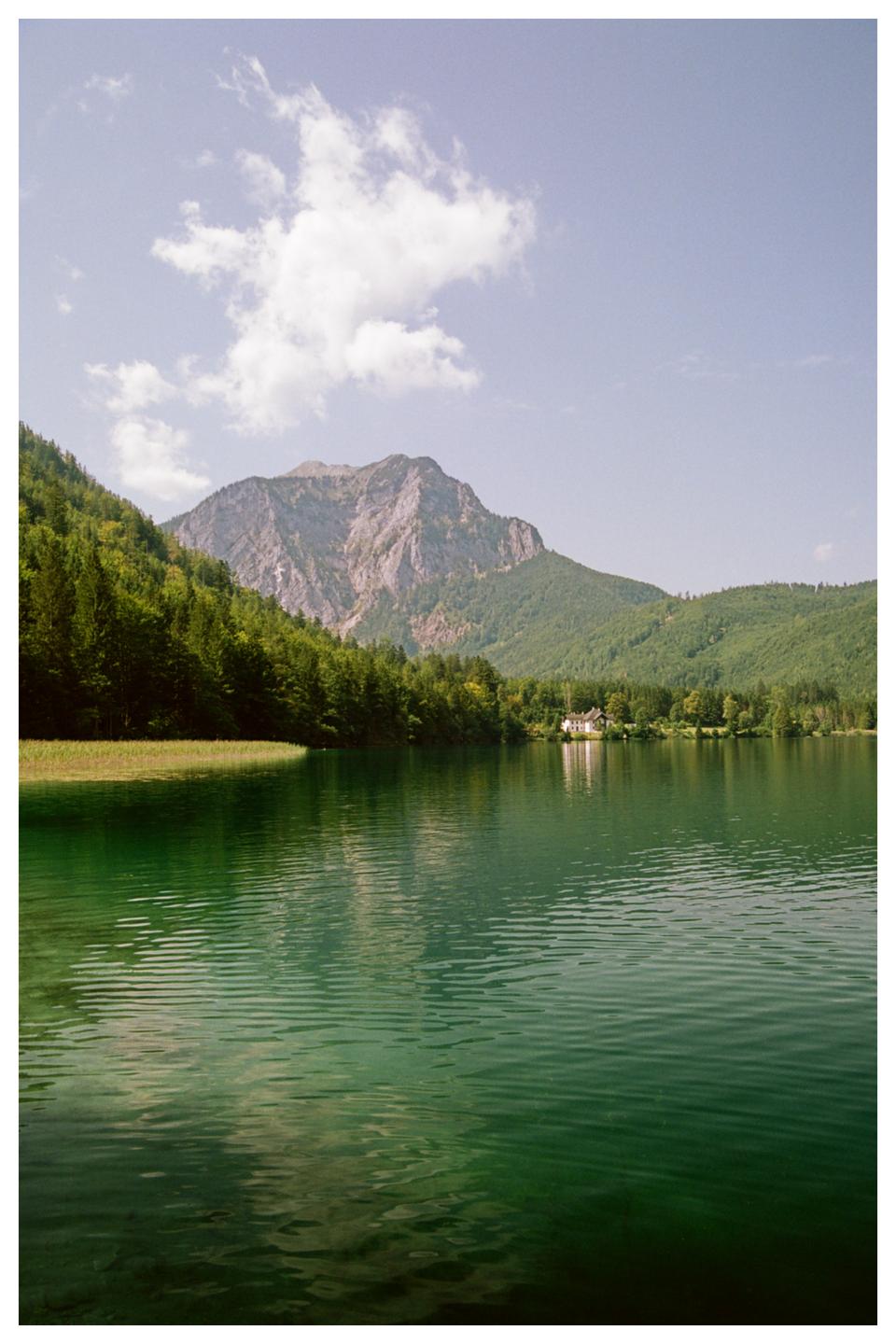 A large body of green water reflects the surrounding forest and mountains under a sky with dispersing clouds.